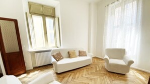 Een ruime woonkamer met een visgraat parketvloer, een witte bank met decoratieve kussens, een witte fauteuil en een groot raam met gordijnen.