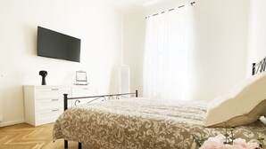 Een slaapkamer met een bed, een nachtkastje met een bloevas, een kaptafel, een aan de muur bevestigde televisie en een raam met gordijnen.
