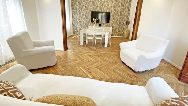 Een woonkamer met een visgraatvloer, witte banken, een eettafel met stoelen en een aan de muur bevestigde televisie.
