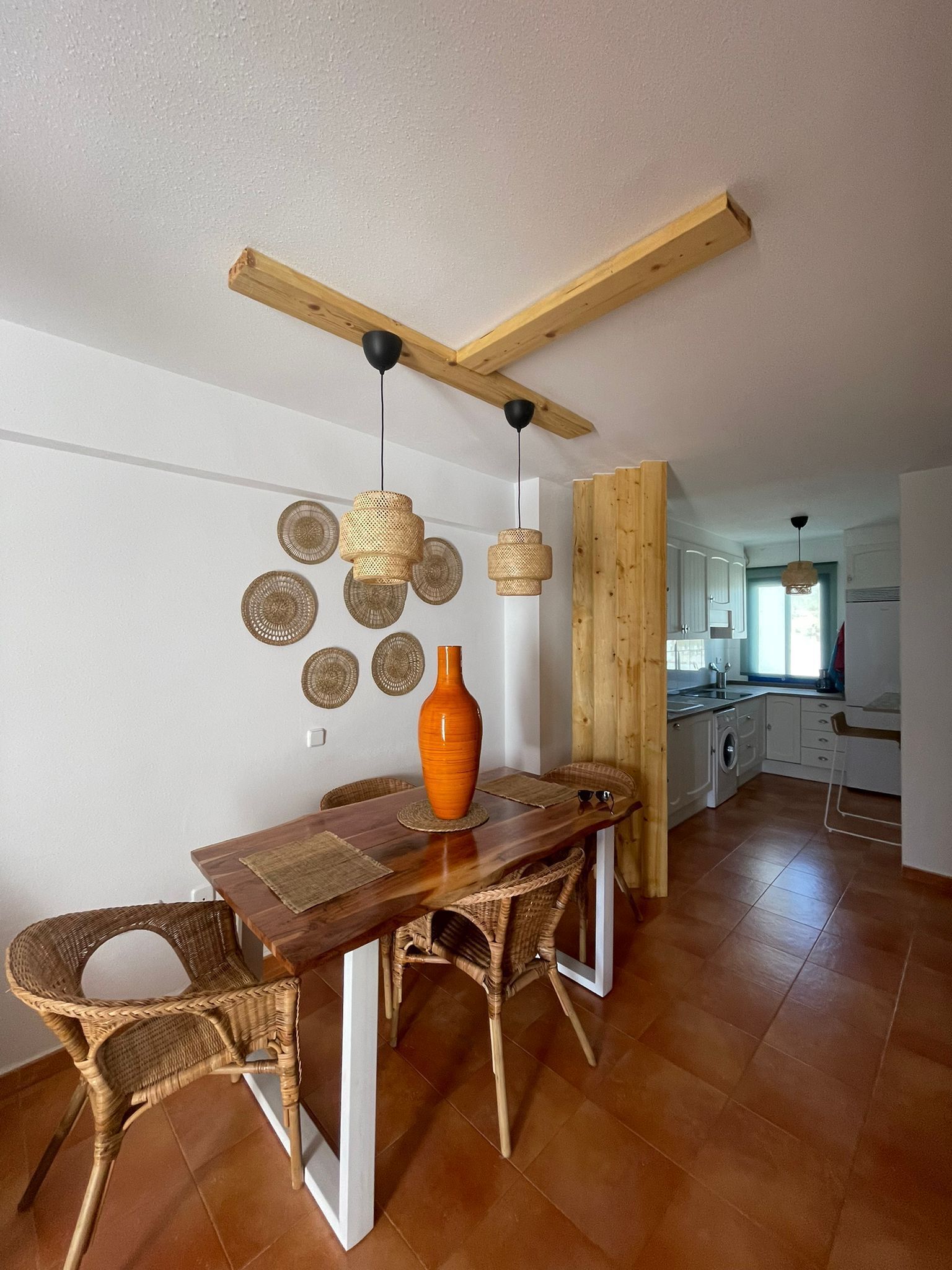 A dining area with a wooden table, wicker chairs, a large orange vase, and woven wall decorations.