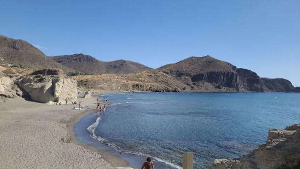 Une plage avec une eau d’un bleu limpide, un rivage sablonneux et des collines rocheuses en arrière-plan.