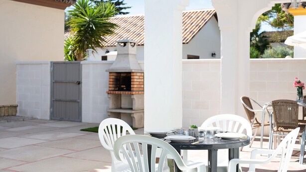 Un patio con sillas blancas y una mesa dispuesta para comer, un horno de ladrillo y vistas a una casa con tejado de baldosas.