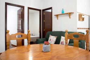 Ein Esstisch mit Stühlen, ein kleiner Topfpflanze, ein grünes Sofa und ein Holzregal in einem Raum mit weißen Wänden und einer Holztür.