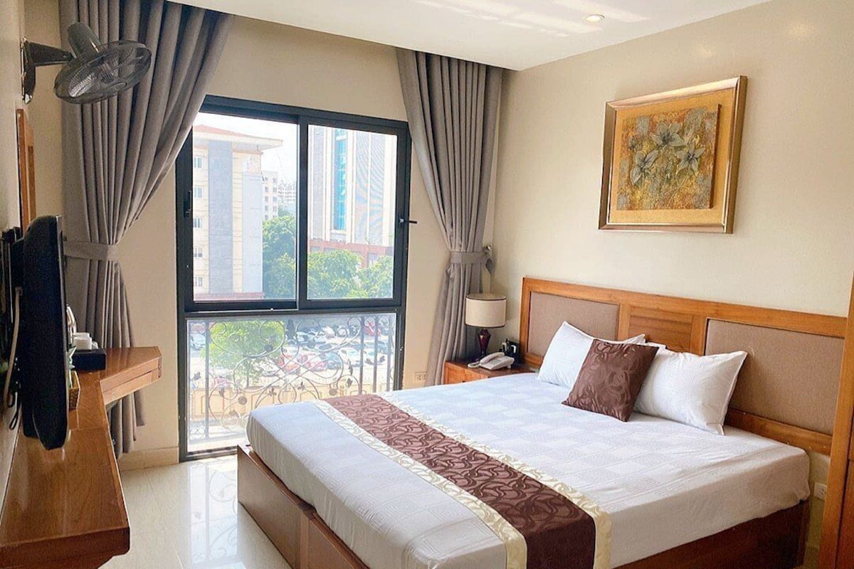 Una habitación de hotel con una cama grande, un cabecero de madera, una mesita de noche con lámpara, una ventana con cortinas y un cuadro en la pared.