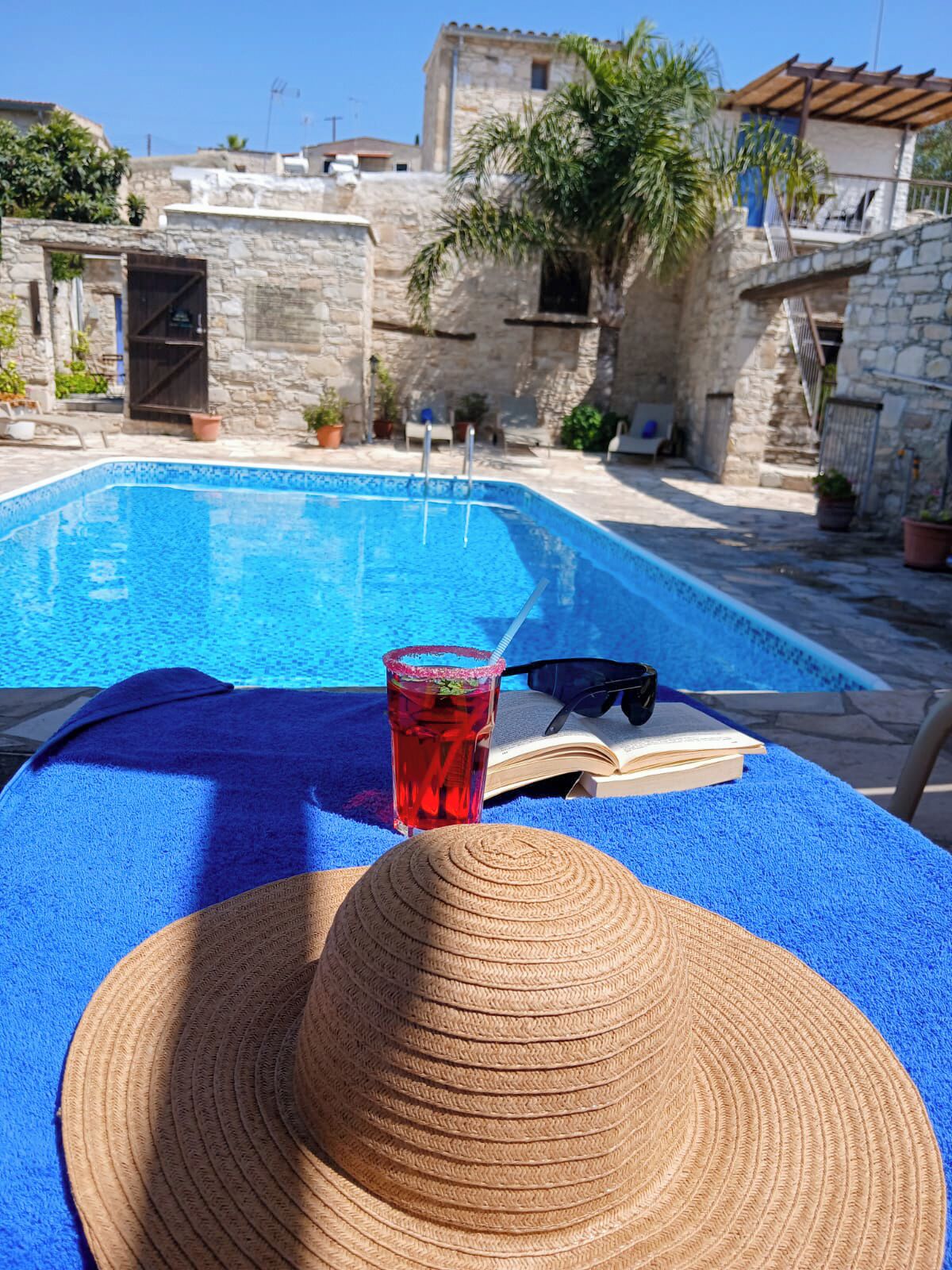 A poolside setting with a blue towel, a straw hat, a drink, and sunglasses on a table.