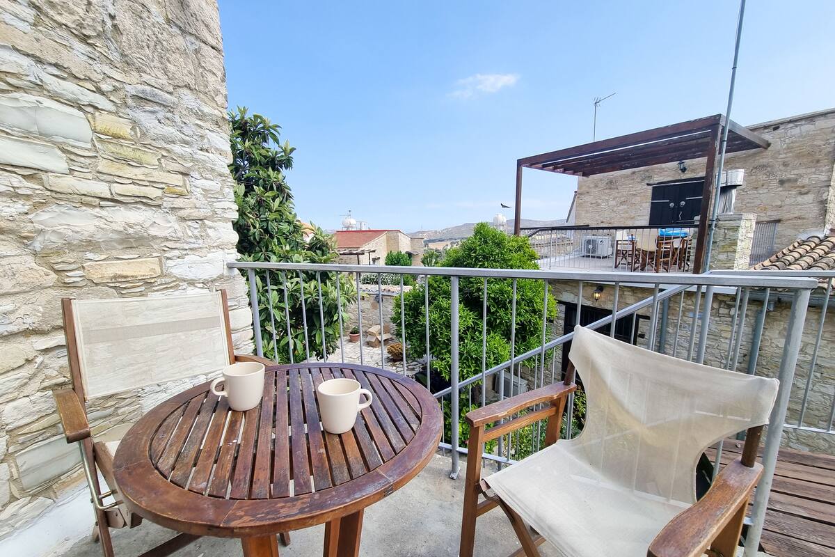 A wooden table with two white cups and a chair with a mesh backrest on a balcony with a stone wall and metal railing.