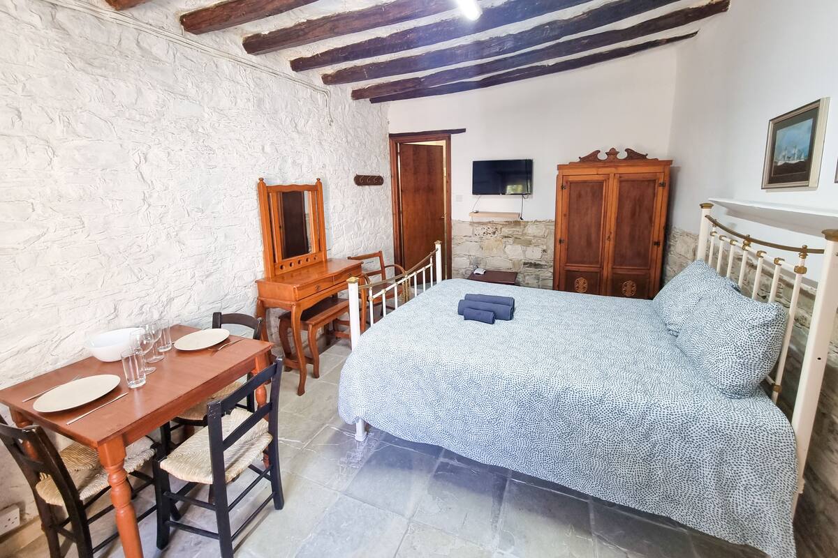 A bedroom with a bed, wooden wardrobe, a desk with a chair, a mirror, and a television on the wall.