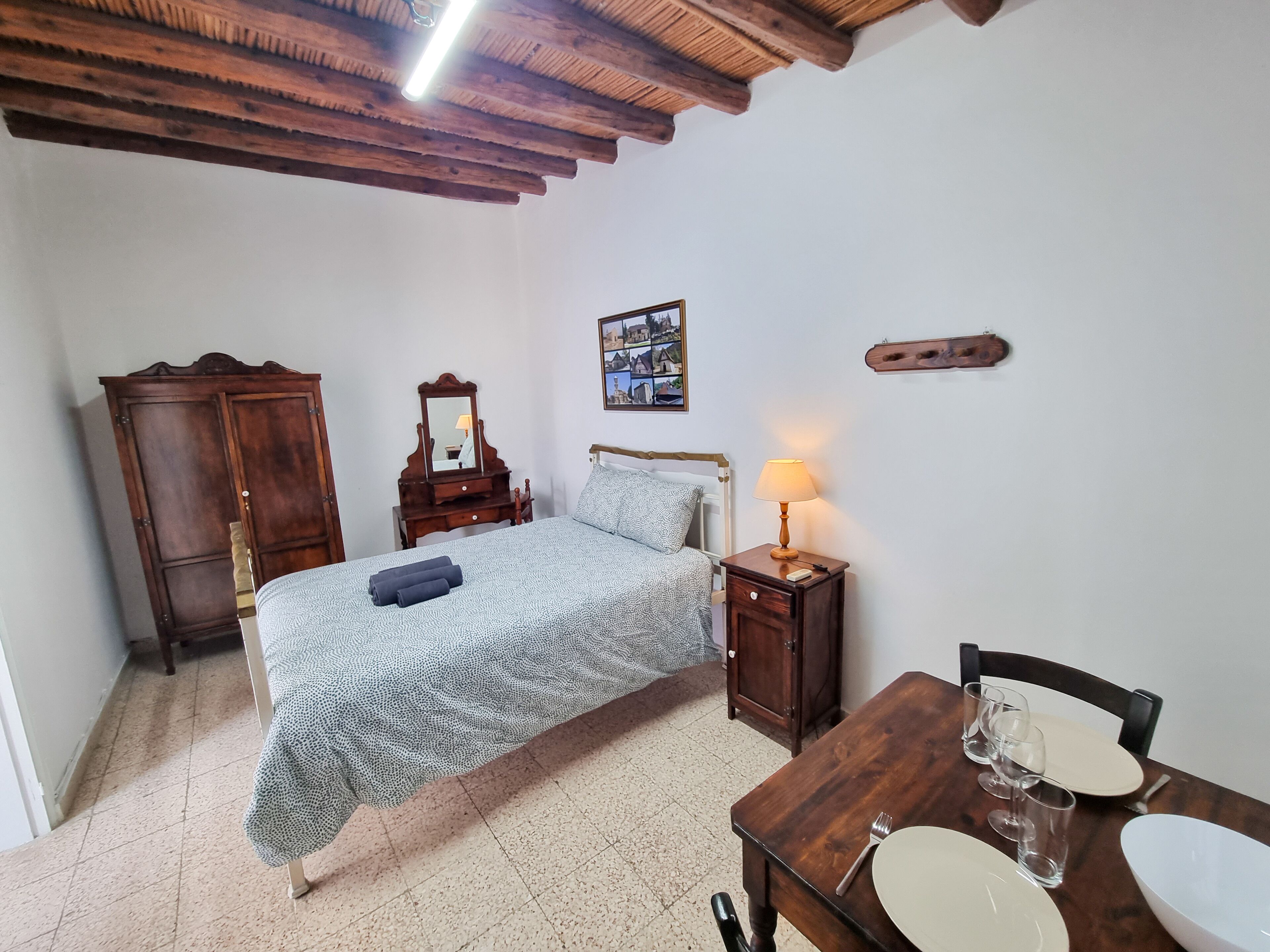 A bedroom with a bed, wooden wardrobe, bedside table with lamp, and a dining table with chairs.