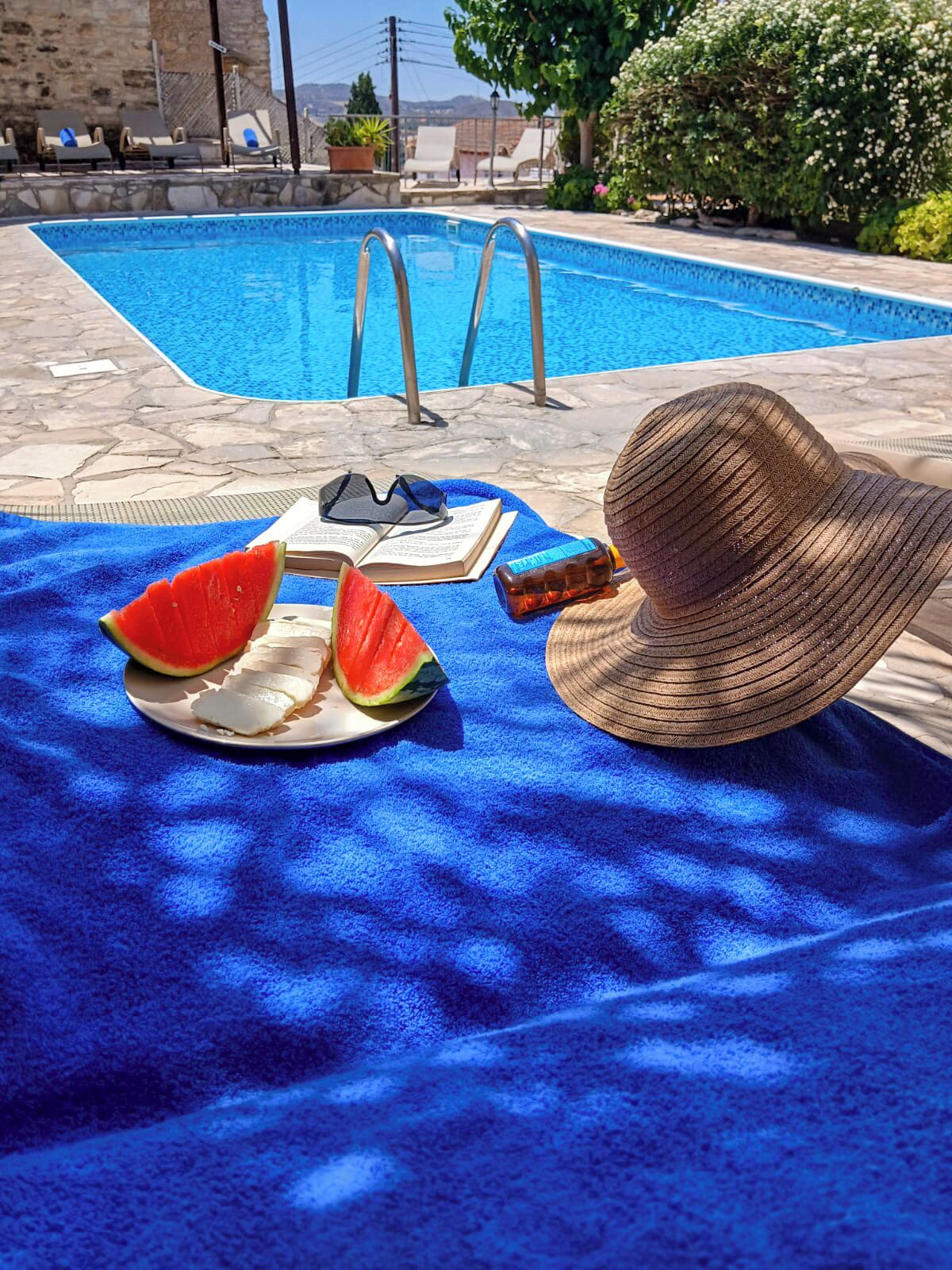A poolside setting with a sun lounger, a hat, watermelon slices, a bottle, and sunglasses on a blue towel.