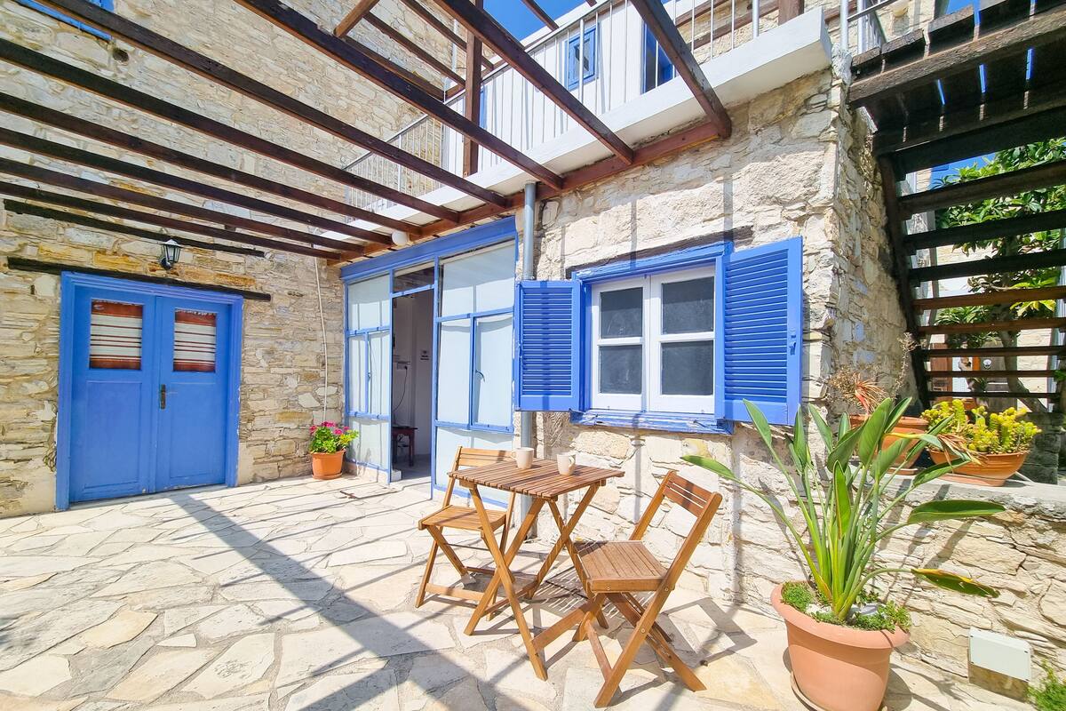 A stone building with a wooden pergola, a blue door, and a table with chairs outside.