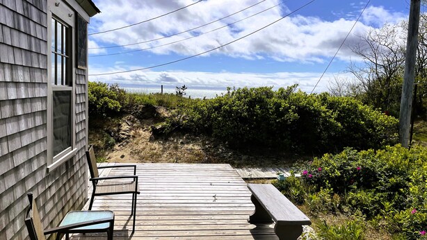 Terraza de madera con un banco y una silla, con vista a una playa con vegetación y un cielo despejado.