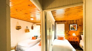 Una habitación acogedora con dos camas, una silla de mimbre, una mesita con lámpara y una ventana con cortinas.