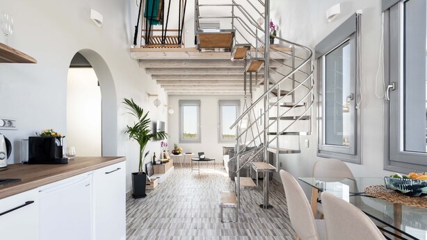 Une cuisine moderne avec des armoires blanches, un plan de travail en bois et un lave-vaisselle encastré. On y trouve un coin repas avec une table et des chaises en verre, ainsi qu’un escalier doté d’une rampe en métal.