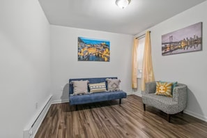 A room with a blue sofa and a patterned armchair, wooden flooring, and two cityscape paintings on the walls.