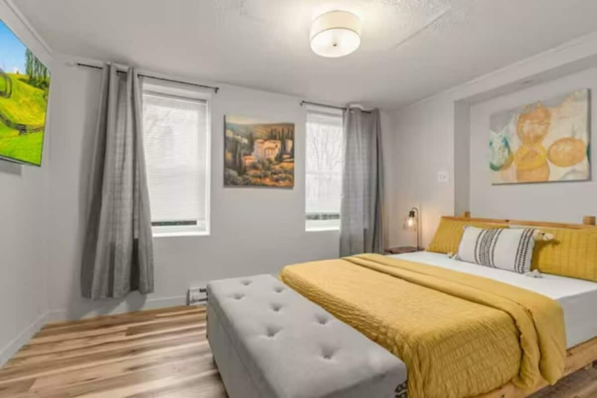 Un dormitorio con cama, una mesita de noche, una ventana con cortinas y un cuadro en la pared.