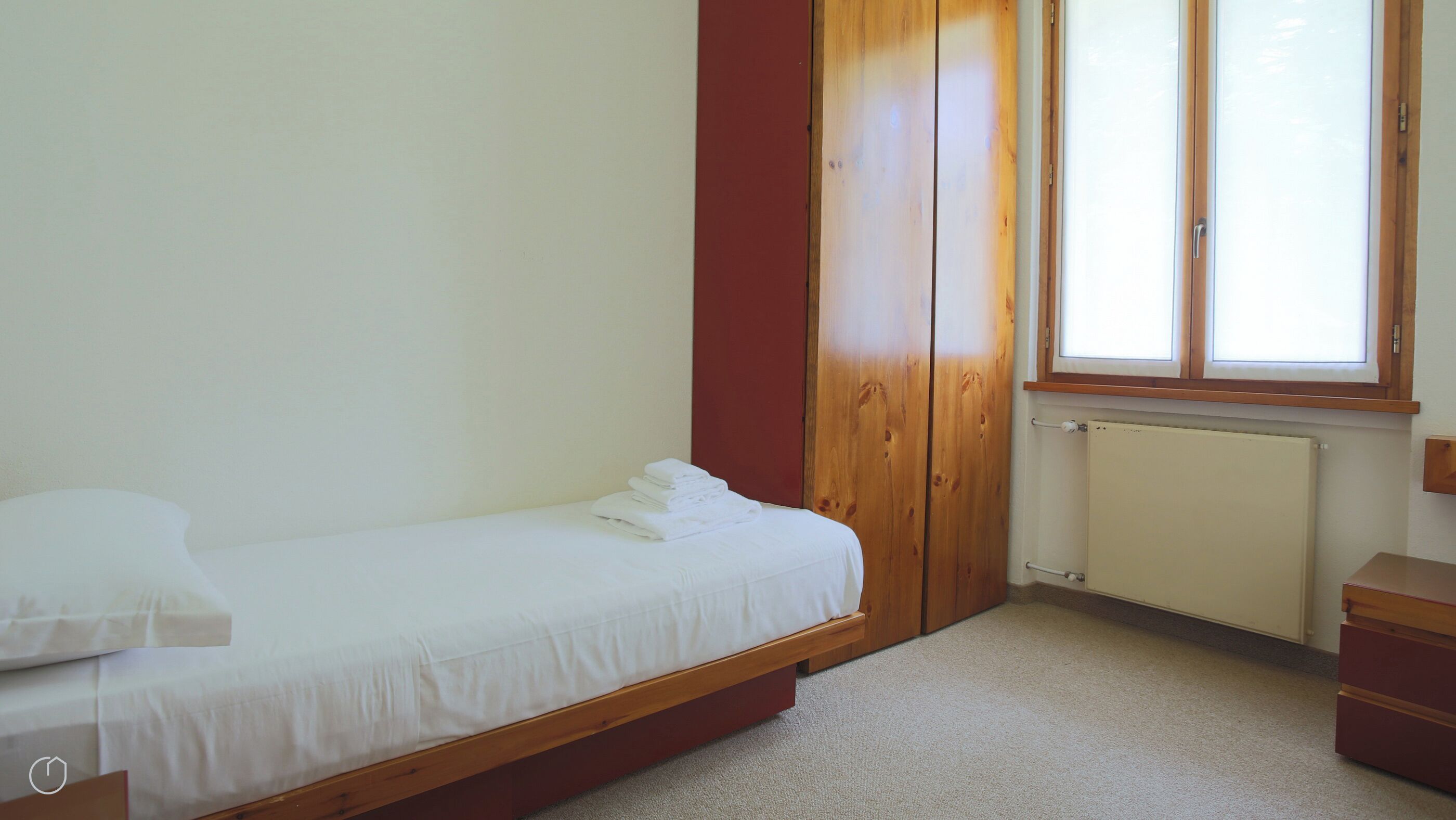 A small, single-bed room with a wooden wardrobe, a radiator, and a window with blinds.