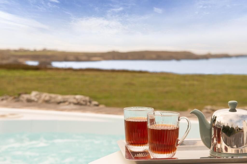 A table with two glasses of tea and a teapot by a poolside with a view of a lake and distant hills.