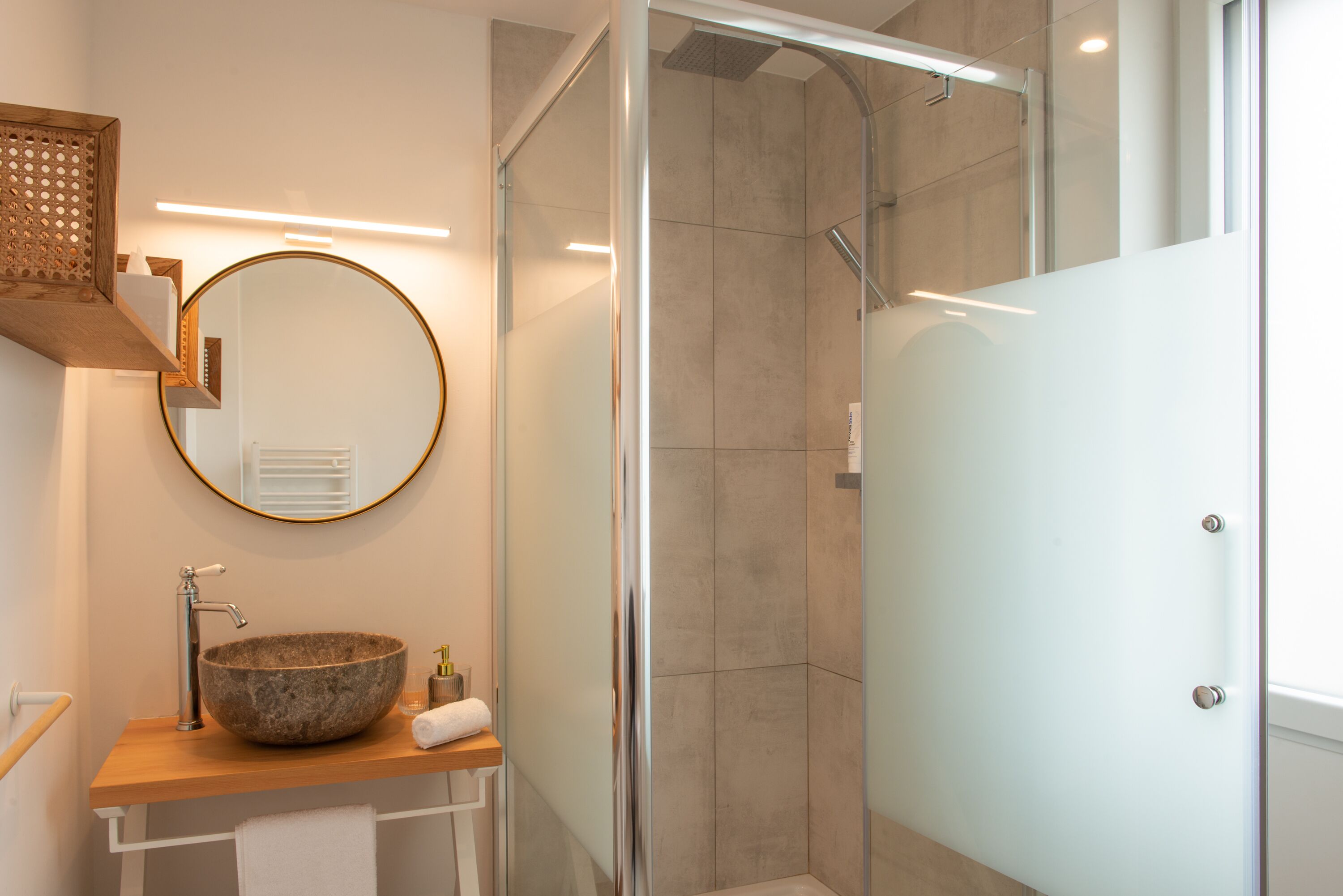 Een moderne badkamer met een ronde spiegel, een wastafel van natuursteen, een douchecabine van glas en een houten plank.