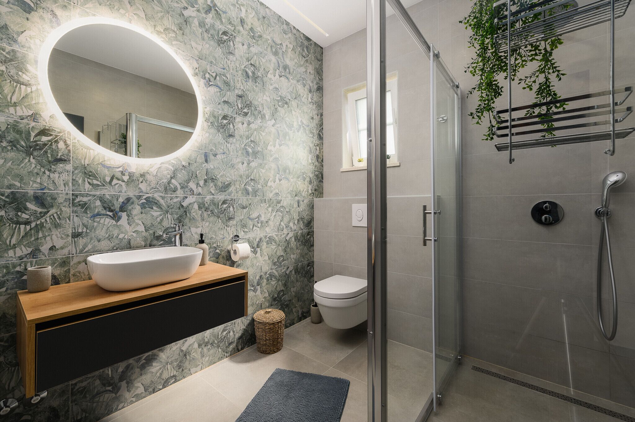 A modern bathroom with a large oval mirror, a white sink, a toilet, and a glass-enclosed shower area.