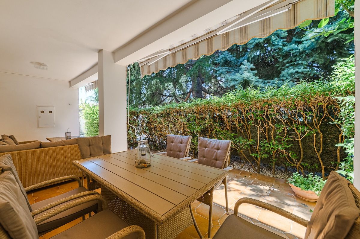 An outdoor dining area with wicker furniture, a wooden table, and a view of a lush garden through a patterned window.