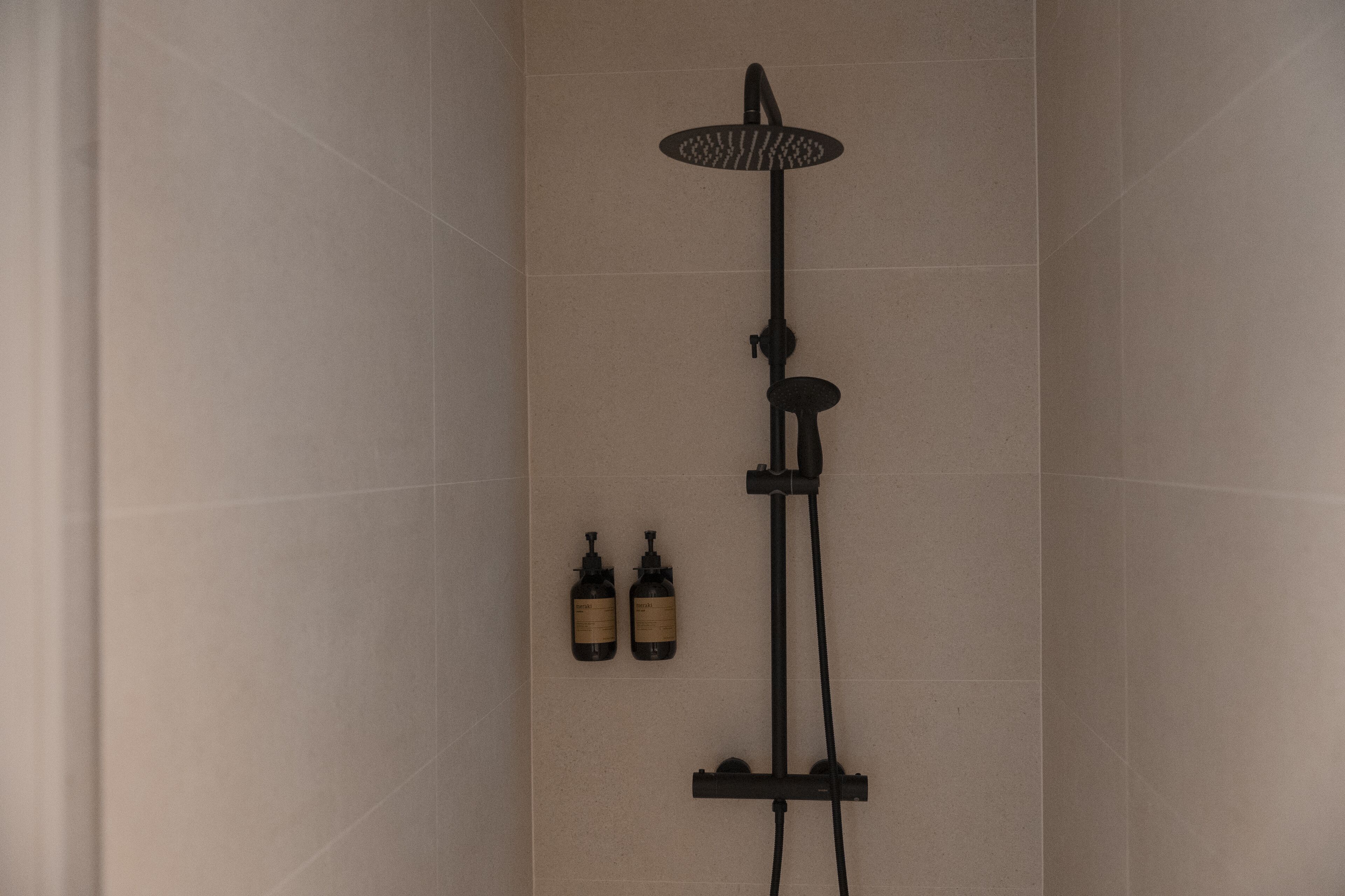 A modern shower with a handheld showerhead and a wall-mounted control panel. There are two bottles of shampoo and conditioner on the wall.