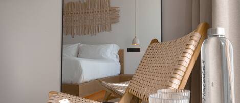 Una habitación de hotel moderna con una cama, una silla de mimbre, una mesita de vidrio y una botella.