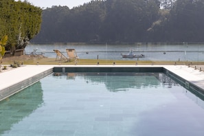 A rectangular pool with clear water, surrounded by a white deck and a row of wooden lounge chairs. A small boat is docked in the background near a forested area. - Malates Slow Boutique Hotel (Foz)