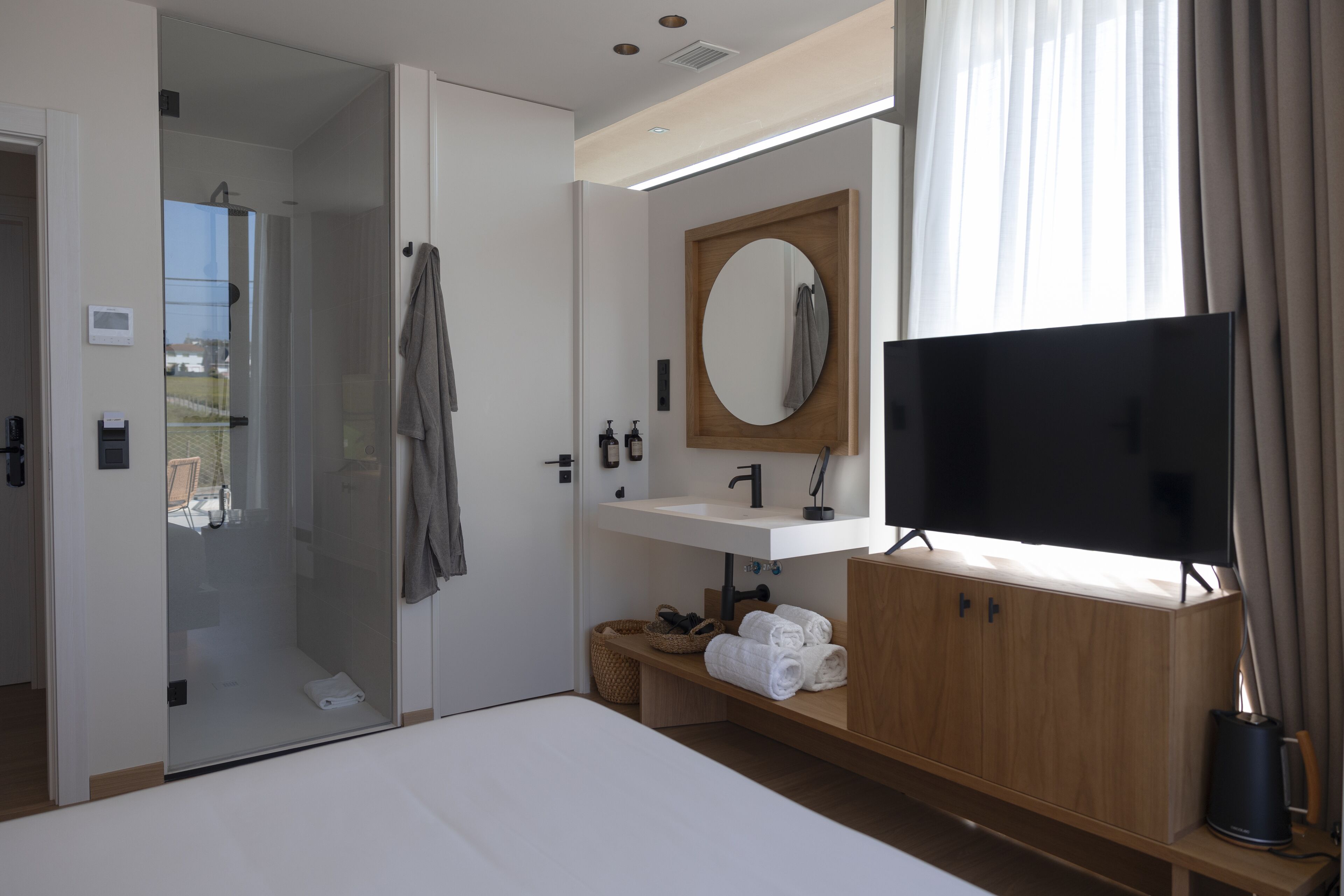Habitación de hotel moderna con ducha de vidrio, tocador de madera con espejo, televisor de pantalla plana y una cama.