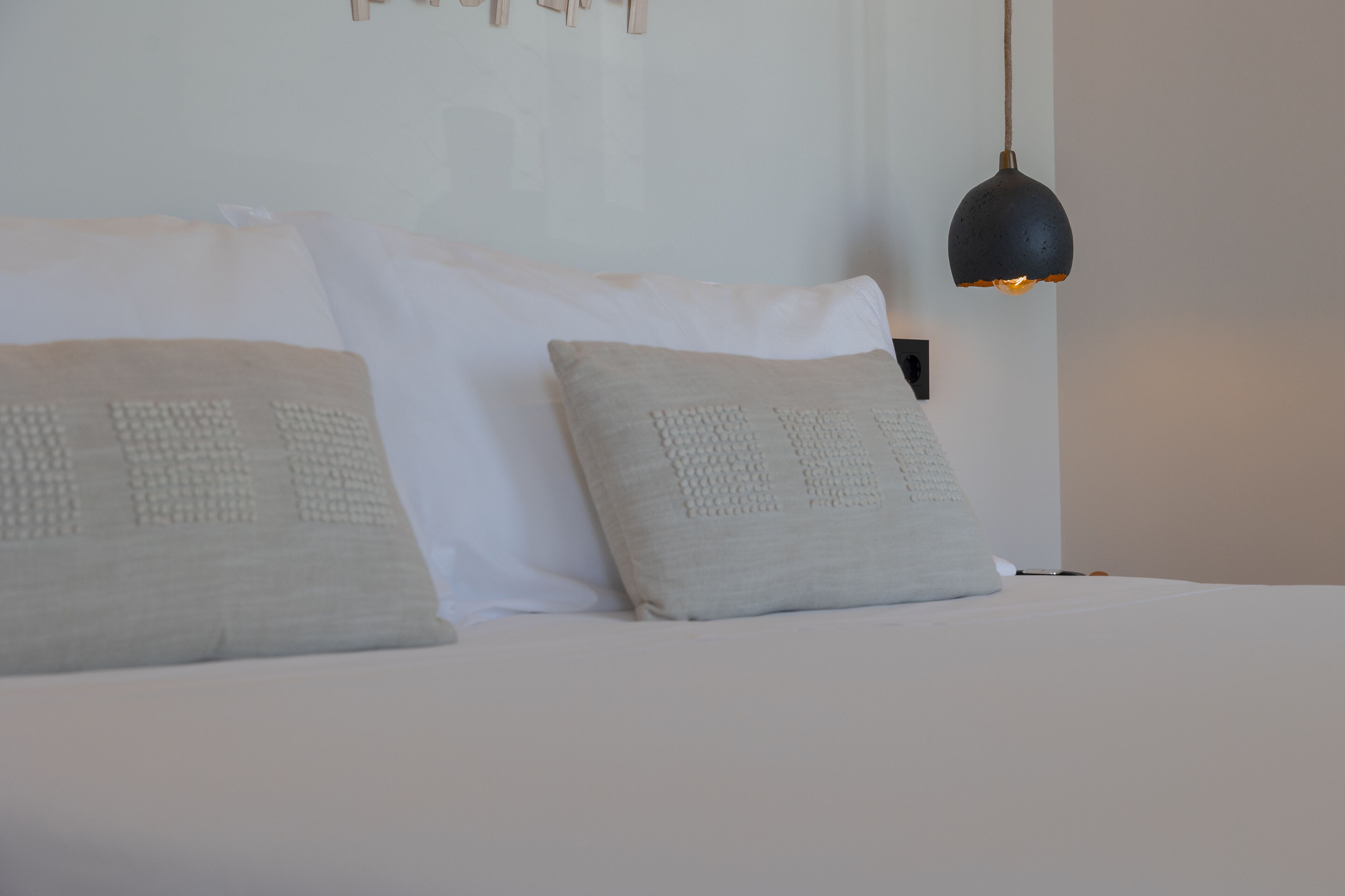 A neatly made bed with white linens and two textured pillows, a black pendant light, and a wall-mounted light switch.