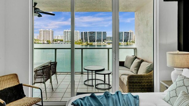Un balcón con vista a un paisaje urbano, una cama con una toalla azul y una silla de mimbre.