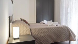 A neatly arranged bedroom with a bed, a bedside table, a wardrobe, and a window with sheer curtains. - La Torre - Deluxe Apartment 1 (Santo Stefano di Camastra)