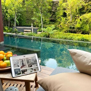 Zona de piscina con una tableta mostrando un anuncio de propiedad, rodeada de exuberante vegetación y una piscina cristalina.