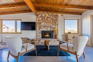 Un salon chaleureux avec une cheminée en pierre, des poutres apparentes en bois et de grandes fenêtres offrant une vue sur les montagnes.