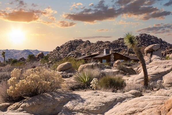 A desert landscape with a wooden cabin surrounded by large rocks and sparse vegetation.