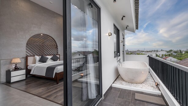 Une chambre moderne dotée d’un grand lit, d’une salle de bain fermée par des parois de verre et agrémentée d’une baignoire sur pieds, et d’un balcon offrant une vue sur la ville.