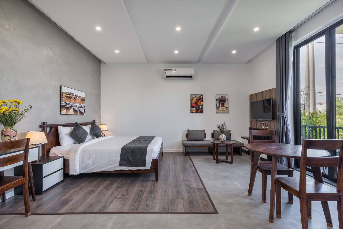 A modern hotel room with a large bed, a sofa, a dining table with chairs, and a TV.