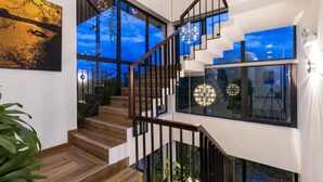 Un escalier moderne avec des rampes en verre et des marches en bois, menant à un deuxième étage doté de grandes fenêtres et d’un luminaire sphérique suspendu.
