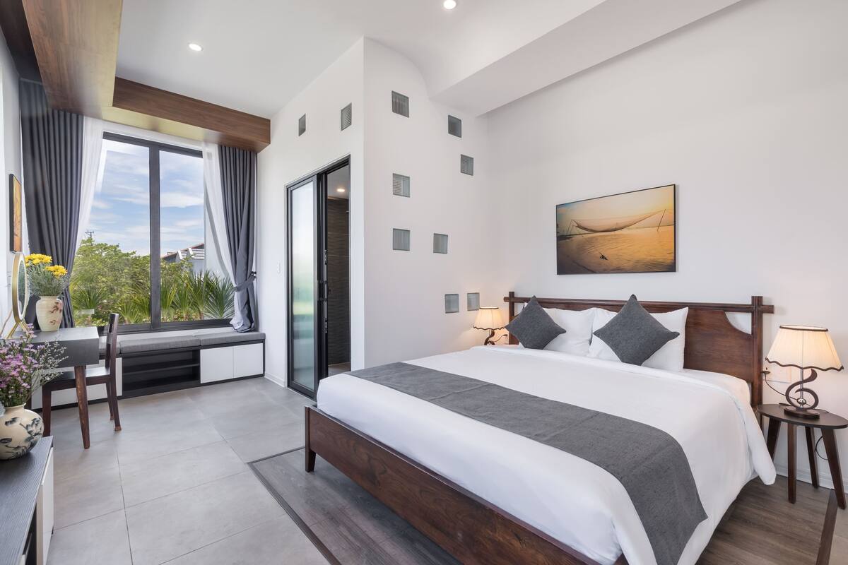 A modern bedroom with a large bed, bedside lamps, a window seat with a table and chairs, and a painting on the wall.