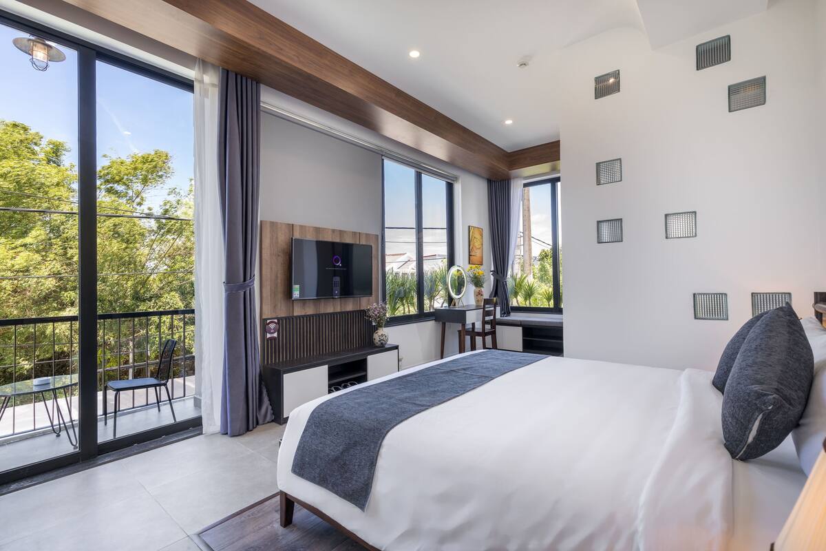A modern bedroom with a large bed, a desk, and a TV. It has a balcony with a view of trees and a building.