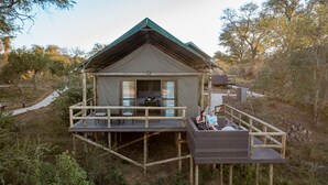 A rustic wooden cabin with a balcony and a covered porch, surrounded by trees and a dirt path.