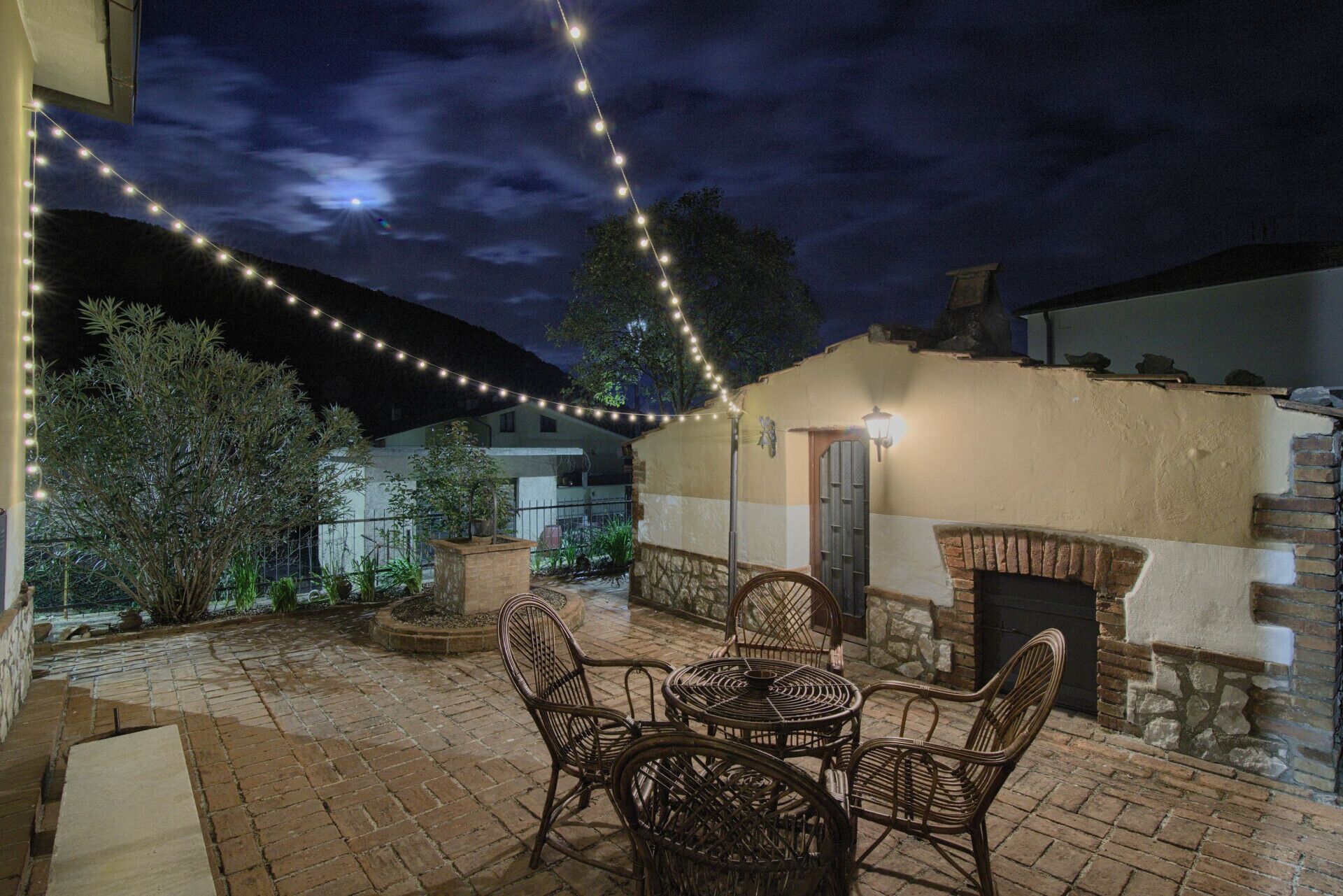Une terrasse extérieure avec des chaises et une table en osier, des guirlandes lumineuses et un mur en pierre agrémenté d’un four en briques.