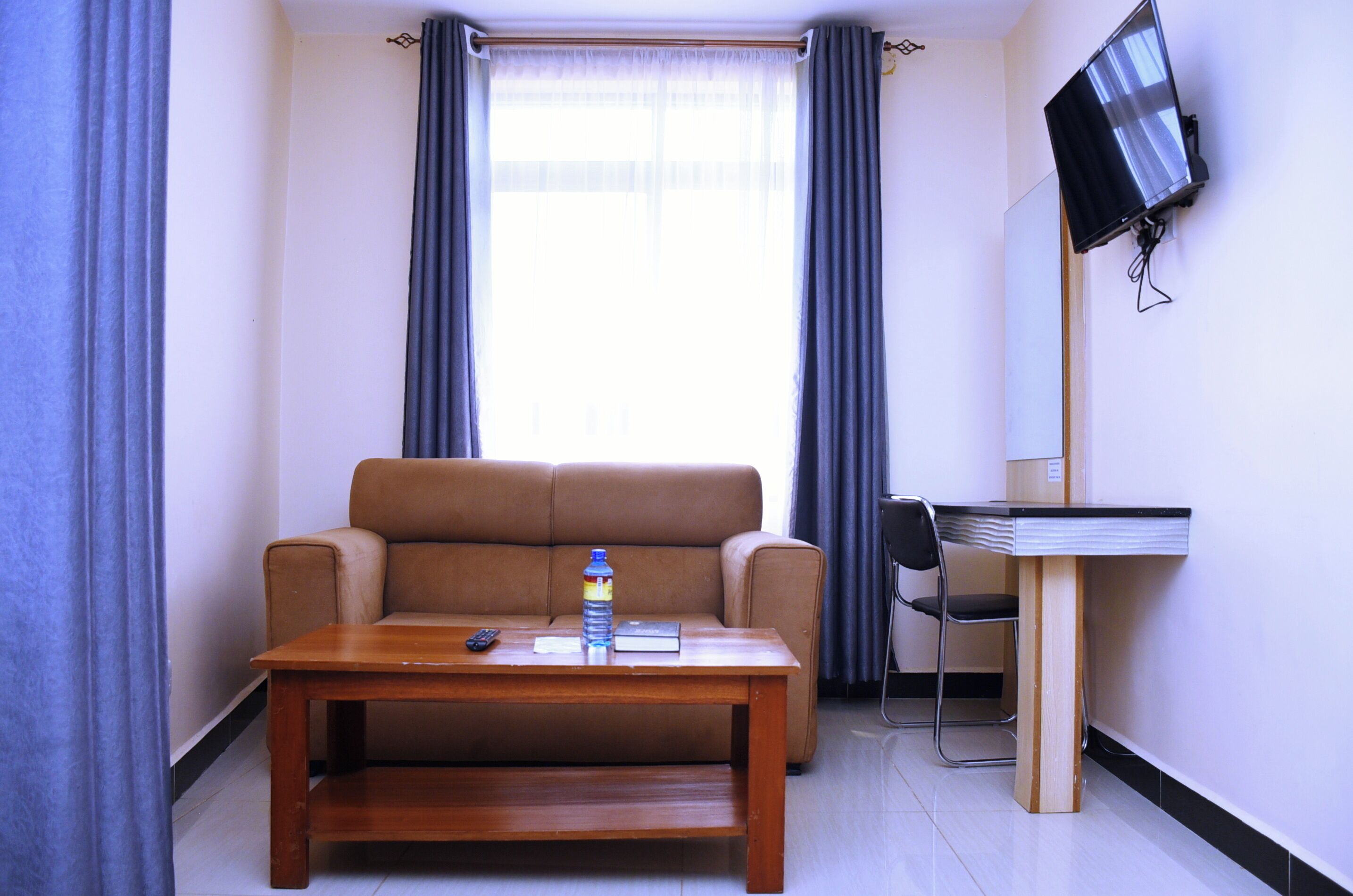 Una habitación pequeña con un sofá, una mesa de madera, una silla, un televisor instalado en la pared y una ventana con cortinas.