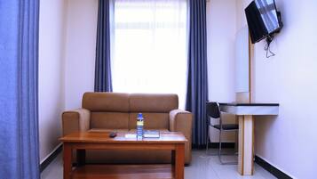 Una habitación pequeña con un sofá, una mesa de madera, una silla, un televisor instalado en la pared y una ventana con cortinas.
