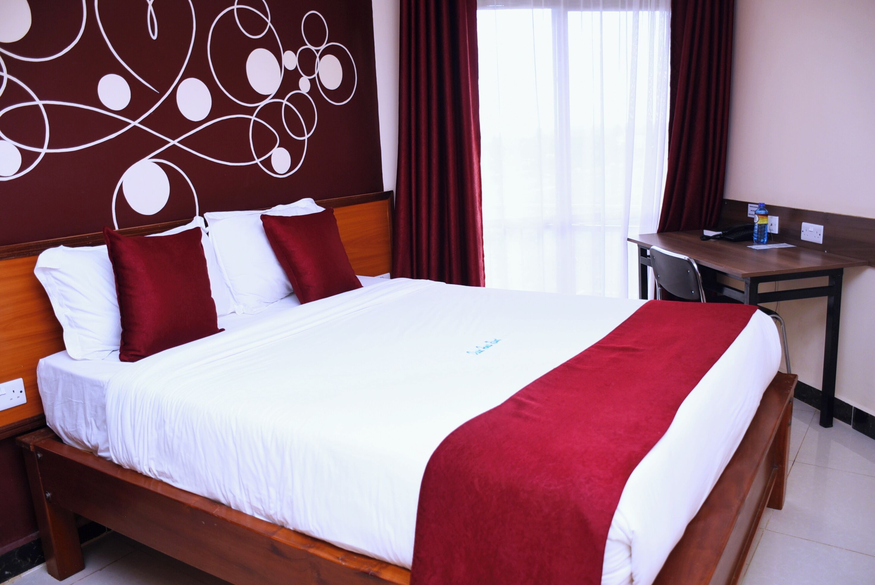 A hotel room with a large bed, red and white bedding, a wooden headboard, a desk with a chair, and a window with red curtains.