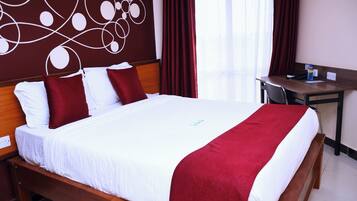 A hotel room with a large bed, red and white bedding, a wooden headboard, a desk with a chair, and a window with red curtains.
