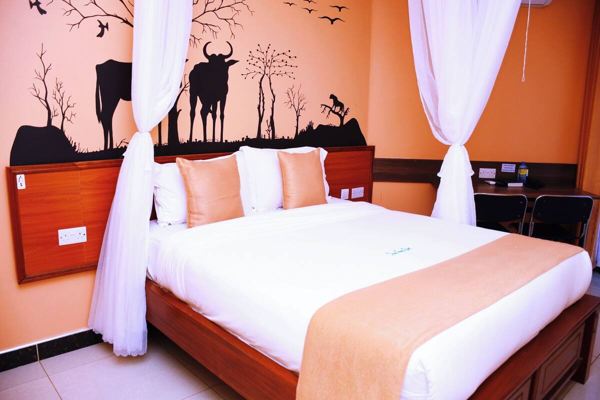 A neatly made bed with white and beige bedding, a wooden headboard, and a wall mural of a rural scene.