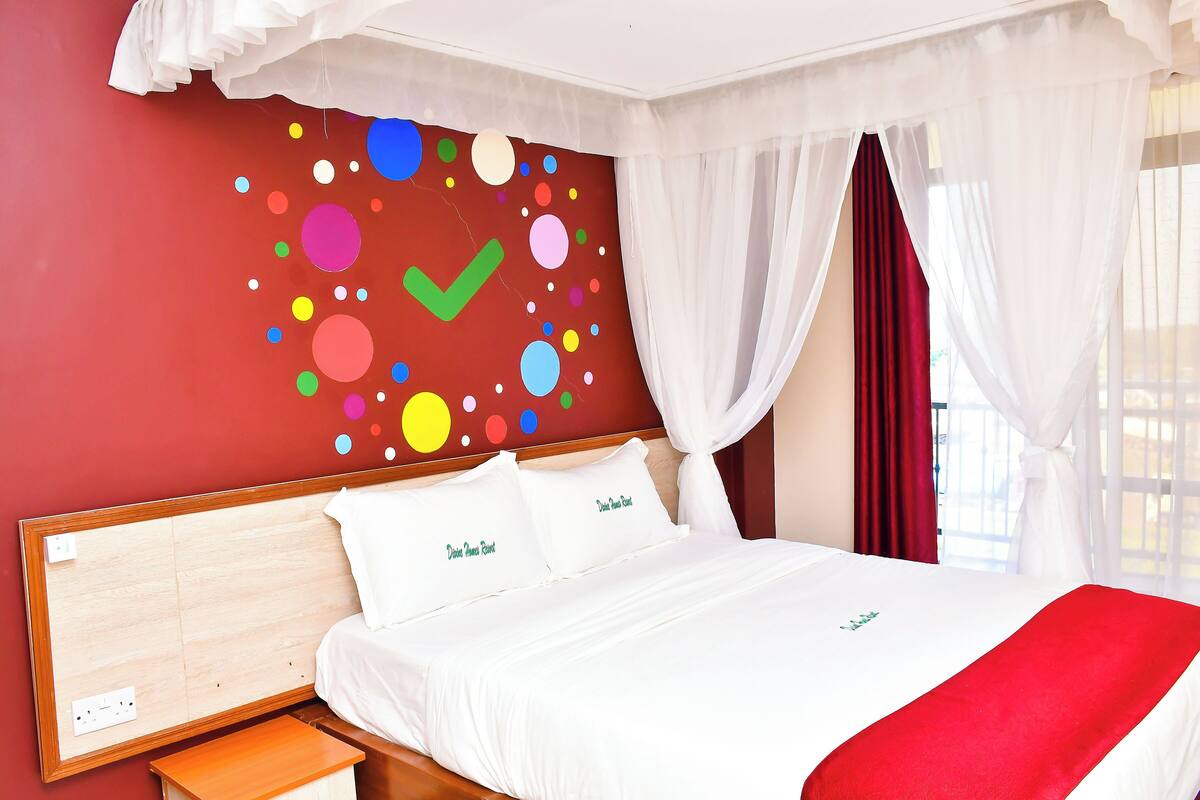 A hotel room with a bed, white bedding, a red wall with colorful circles, and white curtains.