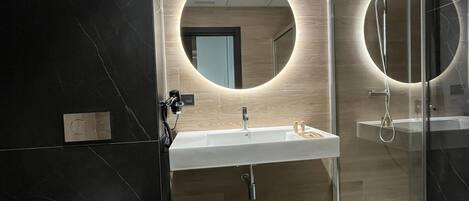 Un baño moderno con un lavamanos blanco, un espejo con luz circular y una cabina de ducha con paredes de vidrio.