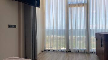 A hotel room with a bed, a TV mounted on the wall, a large window with sheer curtains, and a view of the outdoors.