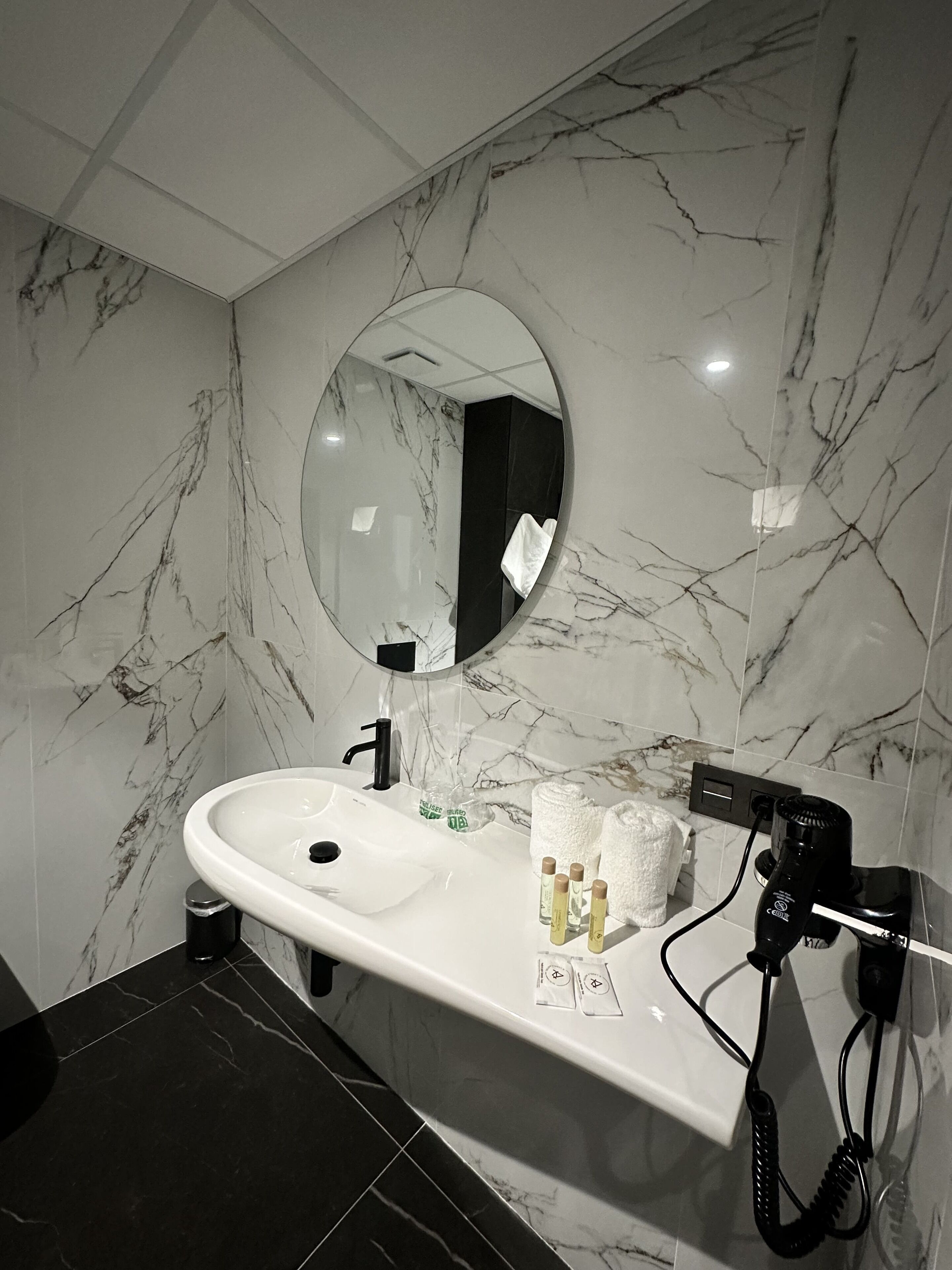 A bathroom with a white sink, a round mirror, and a black hairdryer on the wall.